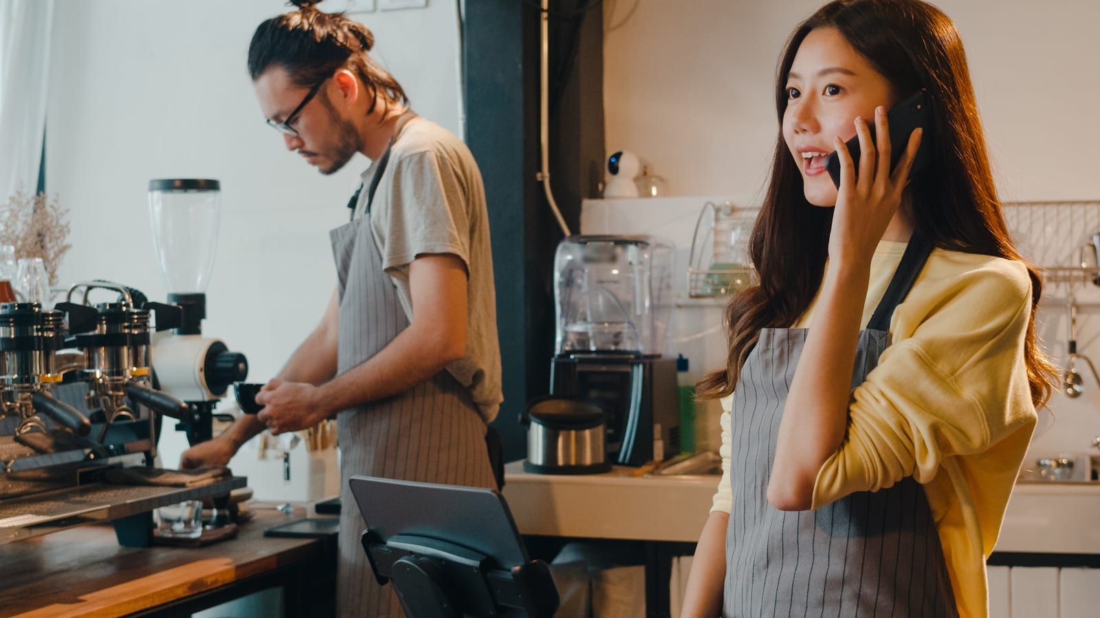 A café worker on the phone beside her till, while a colleague makes coffee behind her