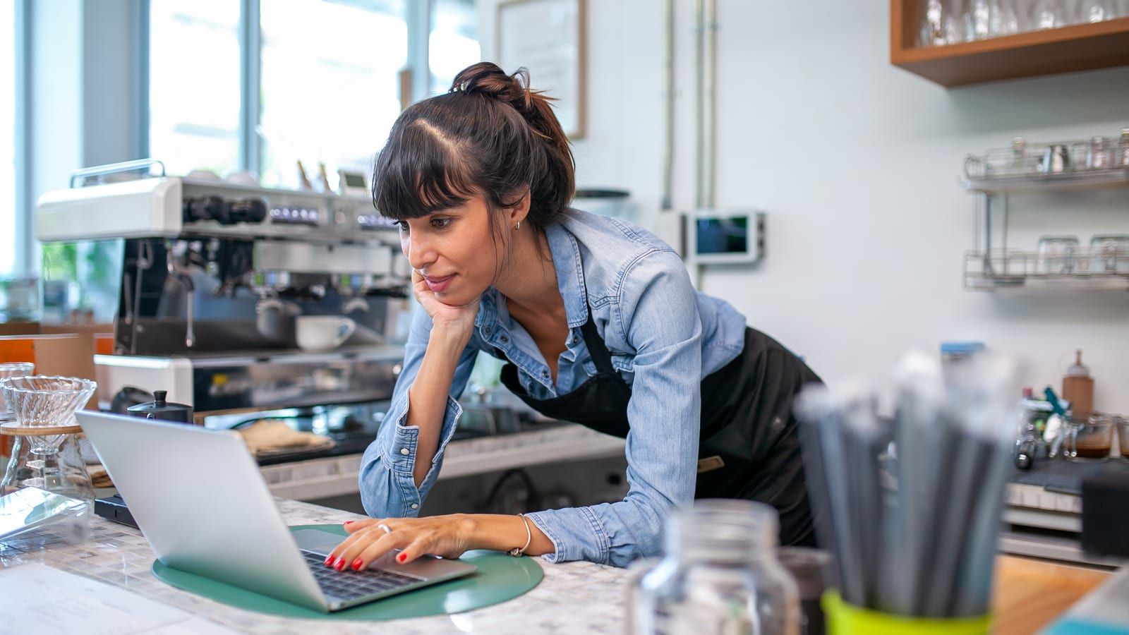 A café owner leaning on the counter, reading something on her laptop